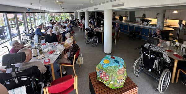 Ældre borgere samles i en lys café til hyggeligt samvær med kaffe, småkager og samtaler ved borde.